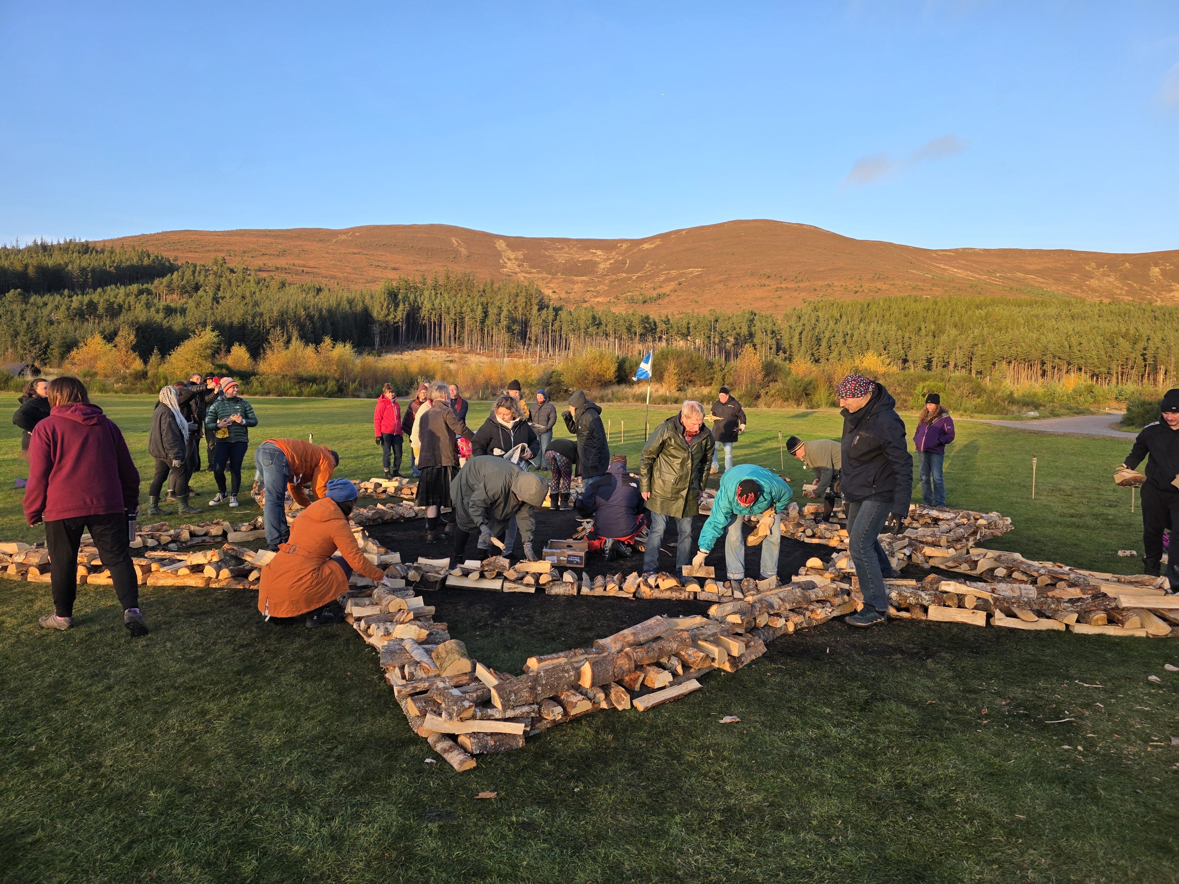 Participants preparing a large outdoor firewalk bed at sunset in the mountains, stacking wood in a star-shaped formation under a clear blue sky surrounded by autumn-colored trees.